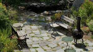 Un patio jardín con un sendero de piedra, un banco de madera, una mesa y una urna decorativa.