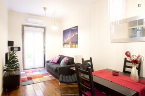 Uma sala de estar com sofá, mesa de jantar com cadeiras, um pequeno pinheiro de Natal e um quadro de uma ponte.