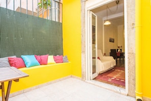 Um quarto amarelo e luminoso com uma porta de vidro deslizante que dá para um quarto. Uma mesa de madeira e almofadas coloridas estão visíveis ao primeiro plano.