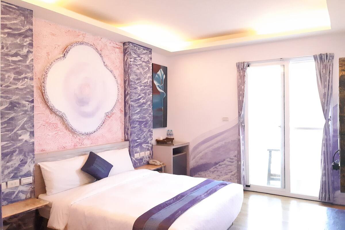 A bedroom with a bed, a nightstand, a wall-mounted phone, and a window with curtains.