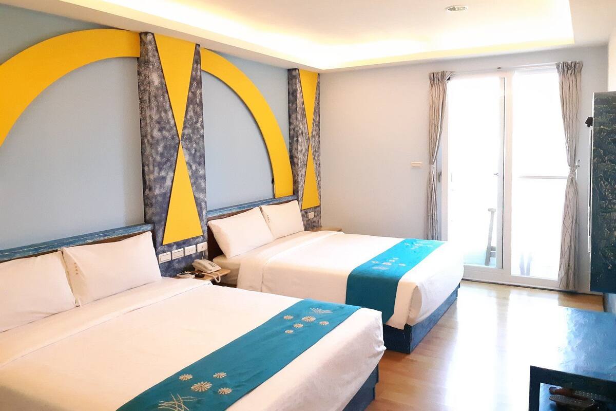 A hotel room with two beds, a desk, and a large window with curtains.