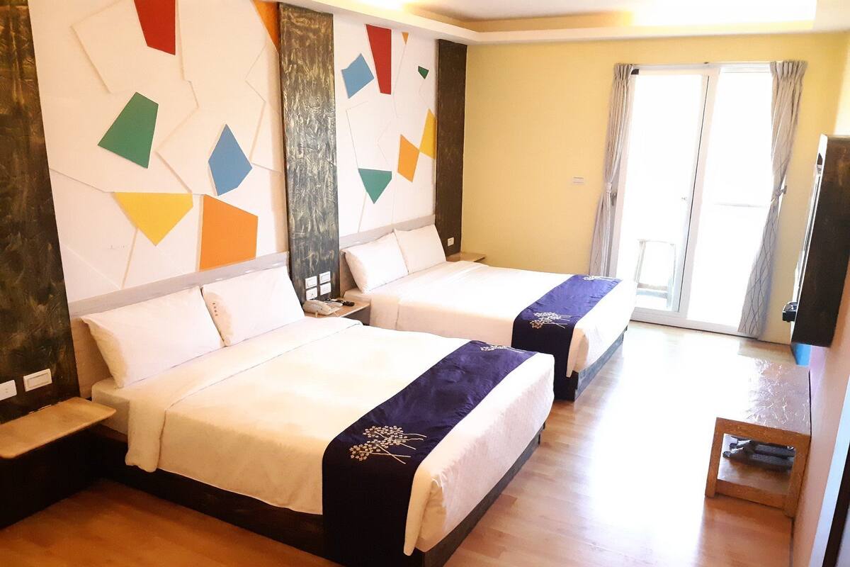 A hotel room with two beds, wooden flooring, a large window with curtains, and a wall decorated with geometric shapes.