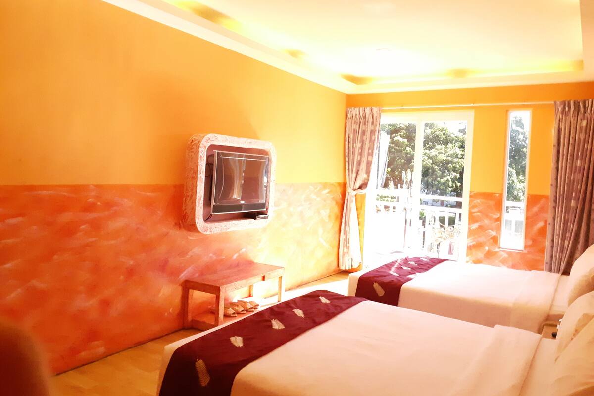 A hotel room with two beds, a TV mounted on the wall, a small wooden table, and a window with curtains.