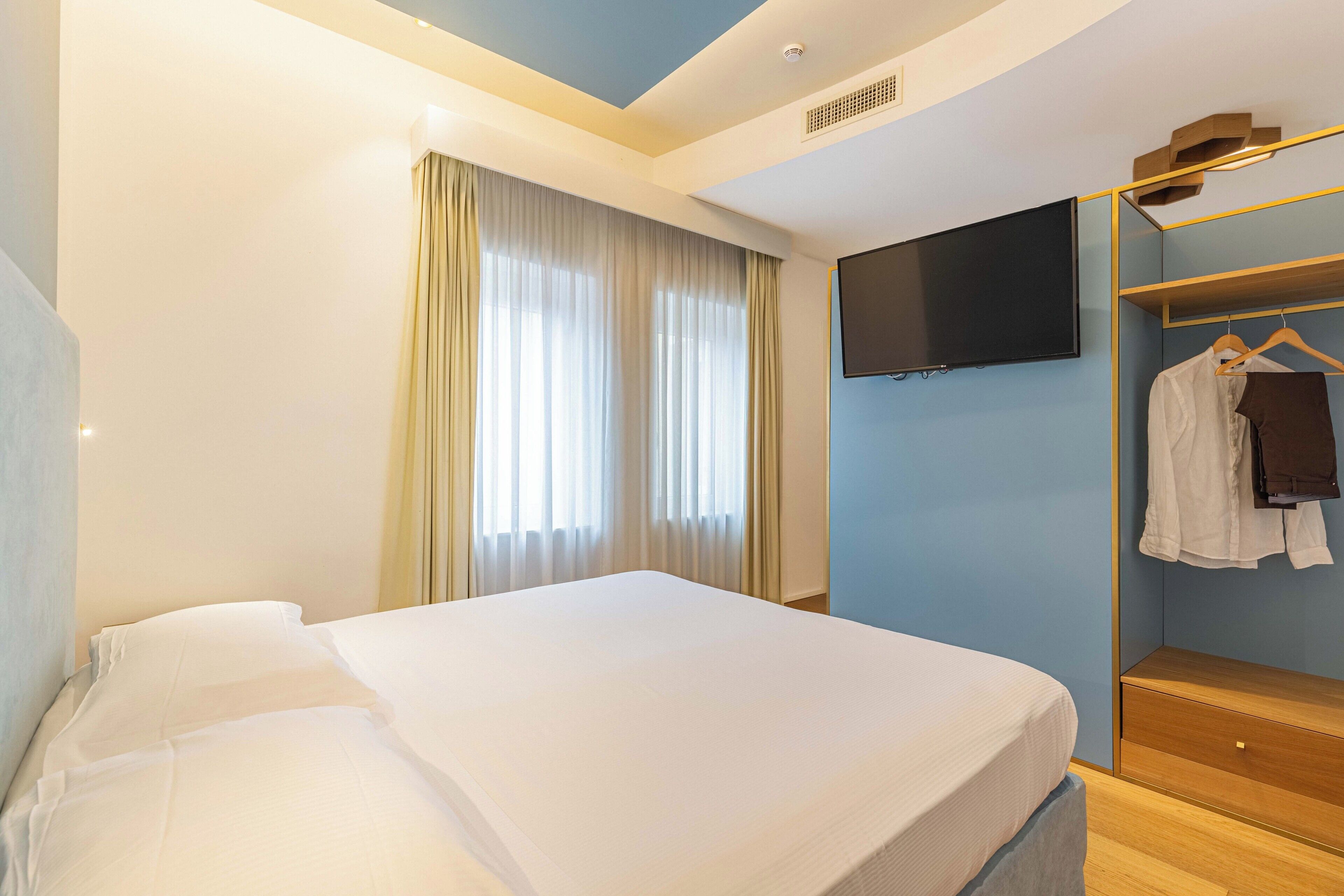 Une chambre d’hôtel avec un lit, un téléviseur fixé au mur, une armoire et une fenêtre avec des rideaux.