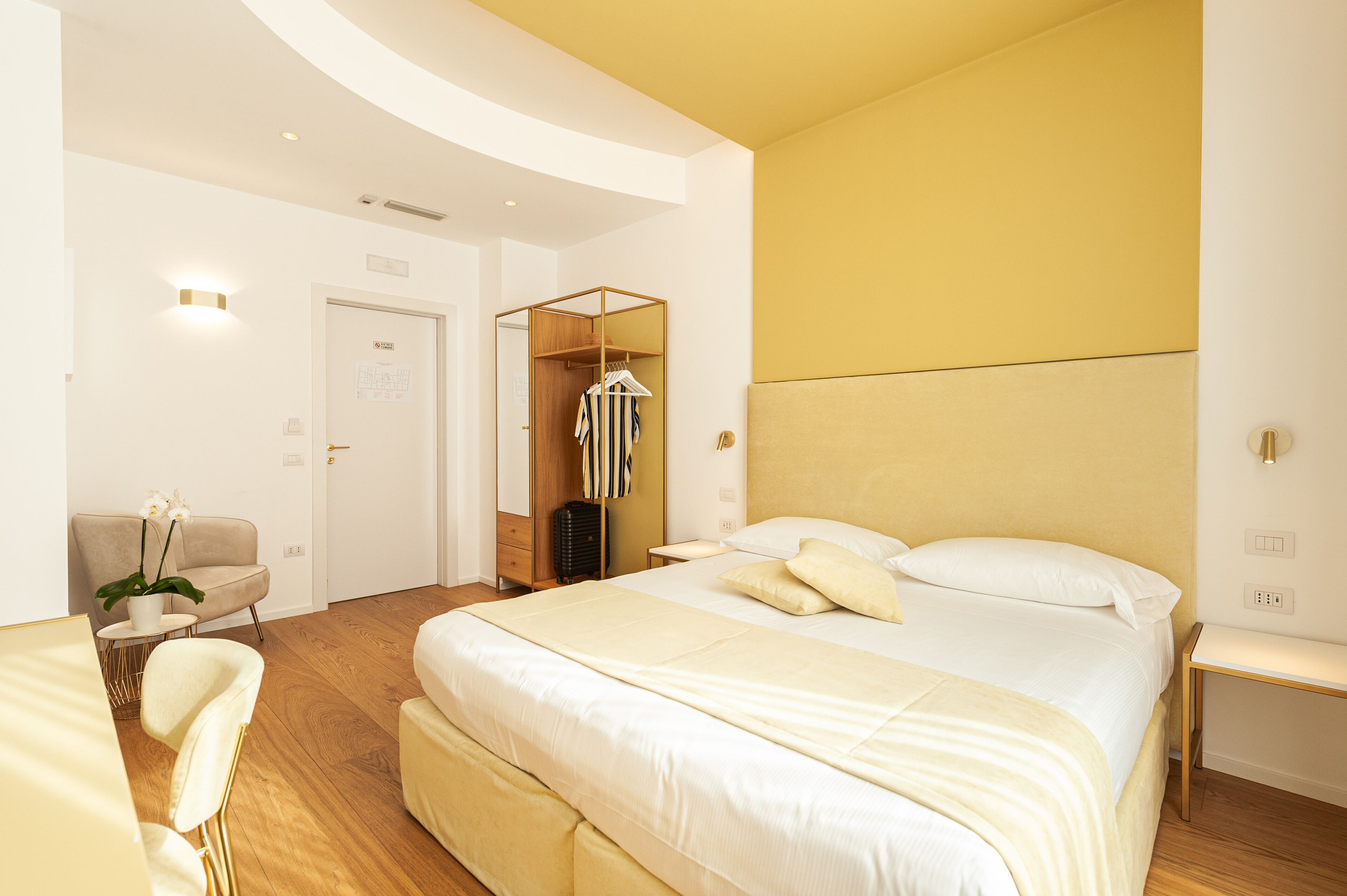 Una habitación de hotel moderna con una cama grande, piso de madera, una zona de estar con una silla y mesa, y un armario con ropa colgada.