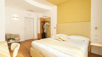 Una habitación de hotel moderna con una cama grande, piso de madera, una zona de estar con una silla y mesa, y un armario con ropa colgada.