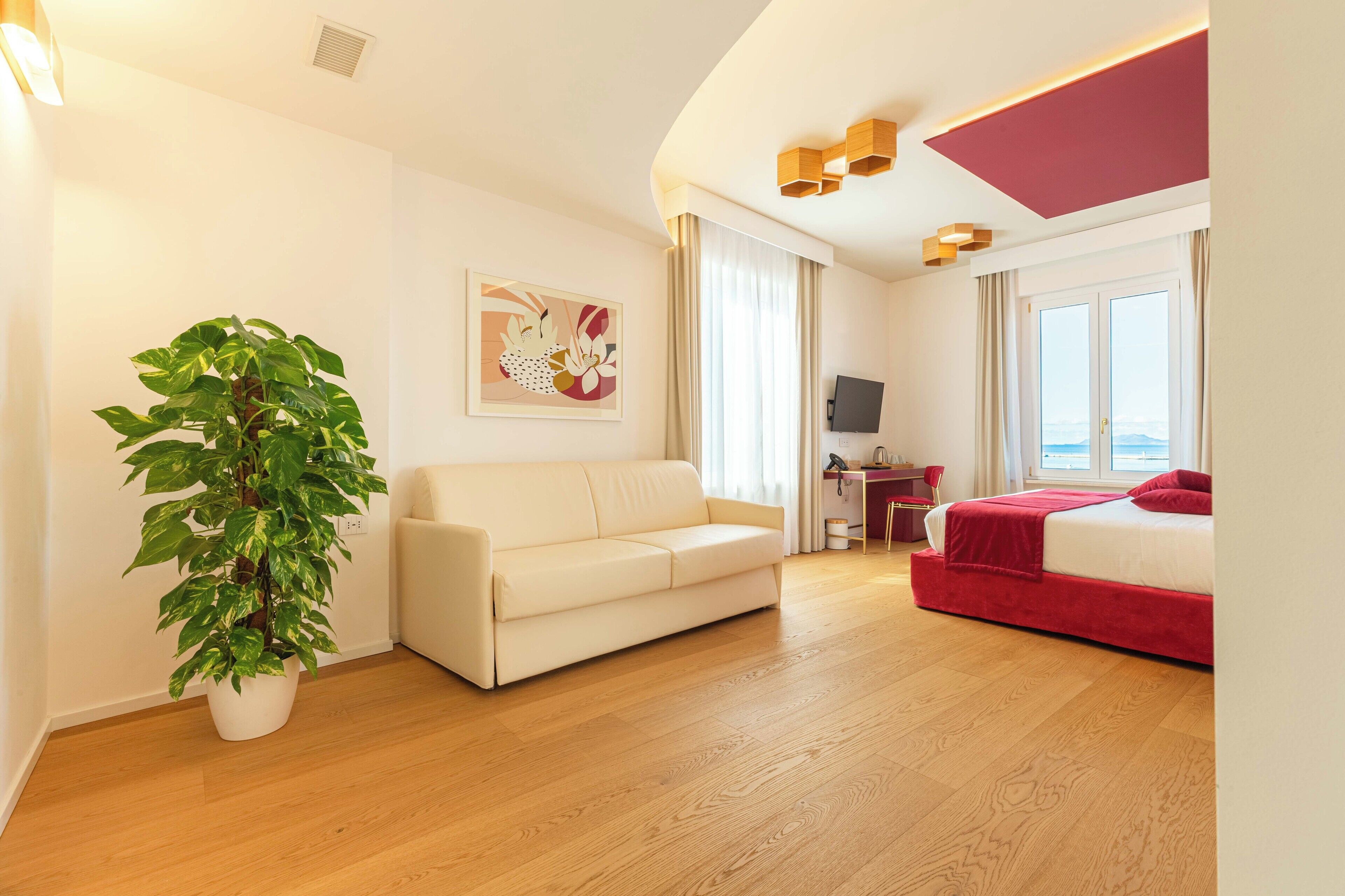 Een moderne hotelkamer met een houten vloer, een witte bank, een rode bed, een grote potplant, een schilderij, een bureau met een computer, en uitzicht op de zee door de ramen.