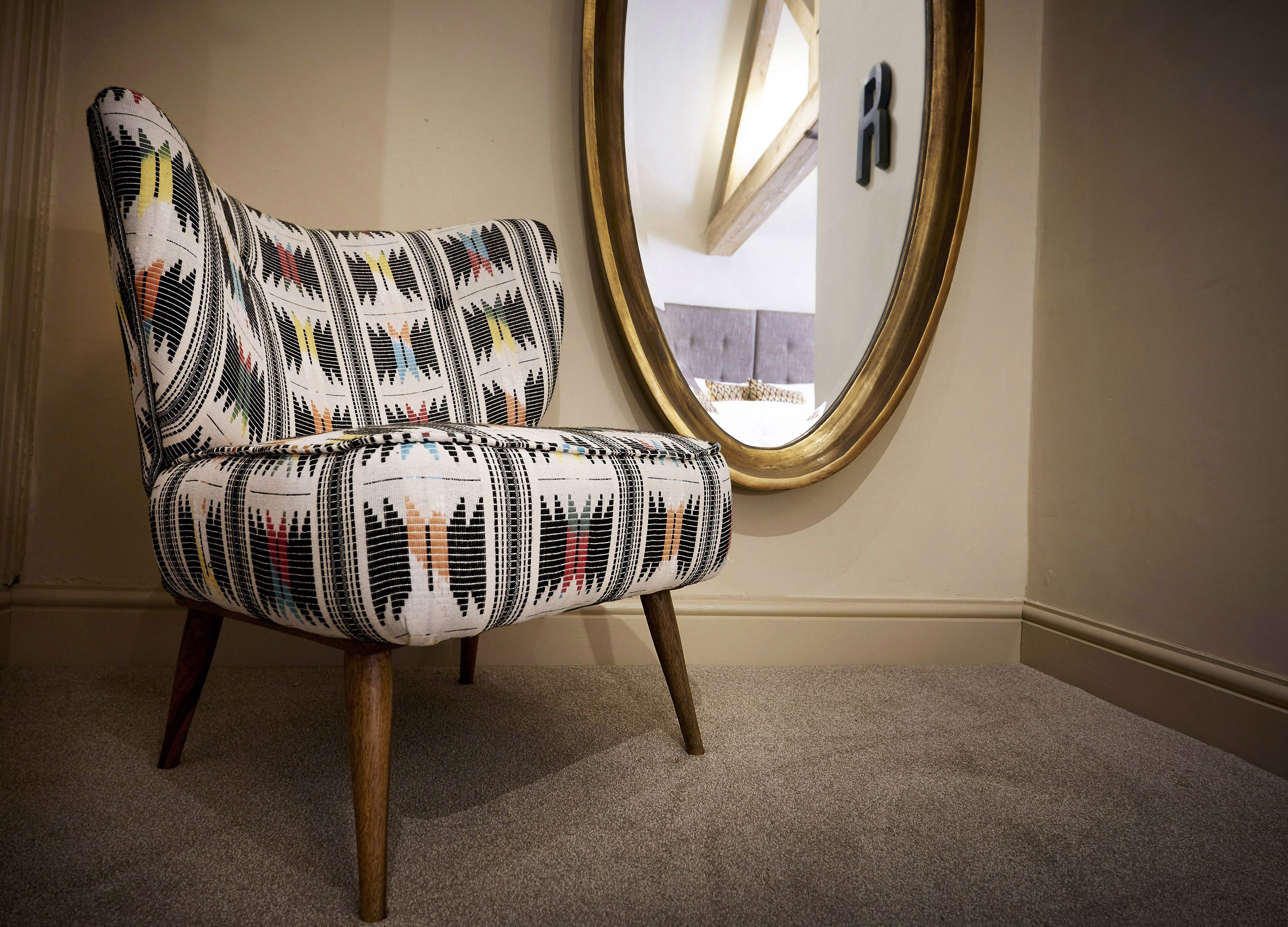 A chair with a patterned fabric and wooden legs in a room with a carpeted floor and a large mirror on the wall.