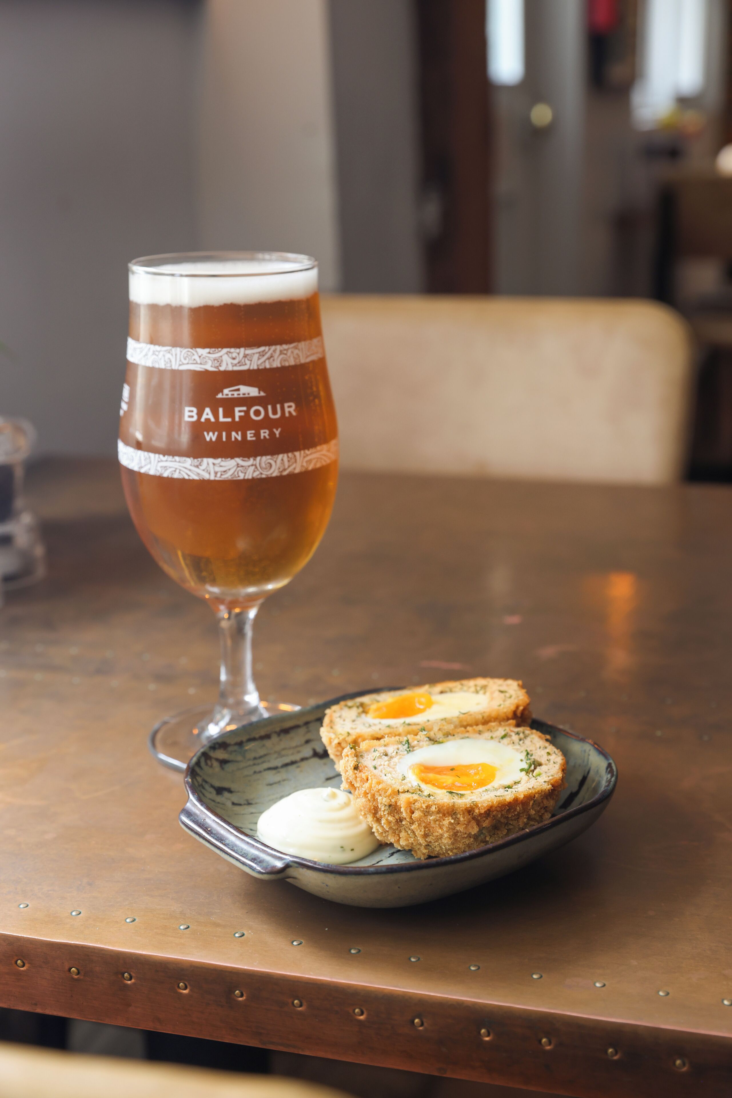 A glass of beer with a label reading "Balfour Winery" and a plate with two pieces of food, possibly Scotch eggs, on a wooden table.