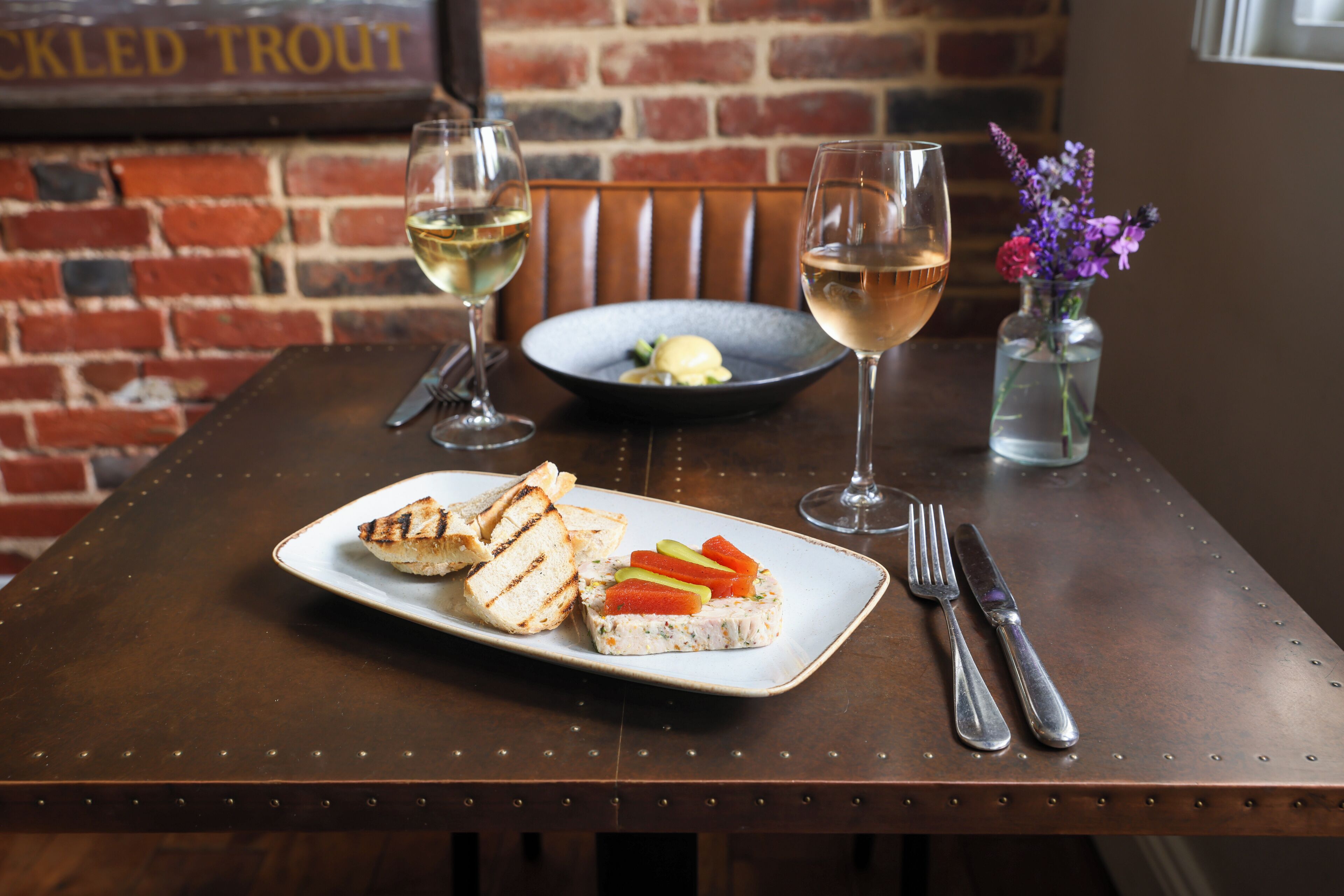 A dining table with a plate of food, two glasses of white wine, and a vase with flowers.