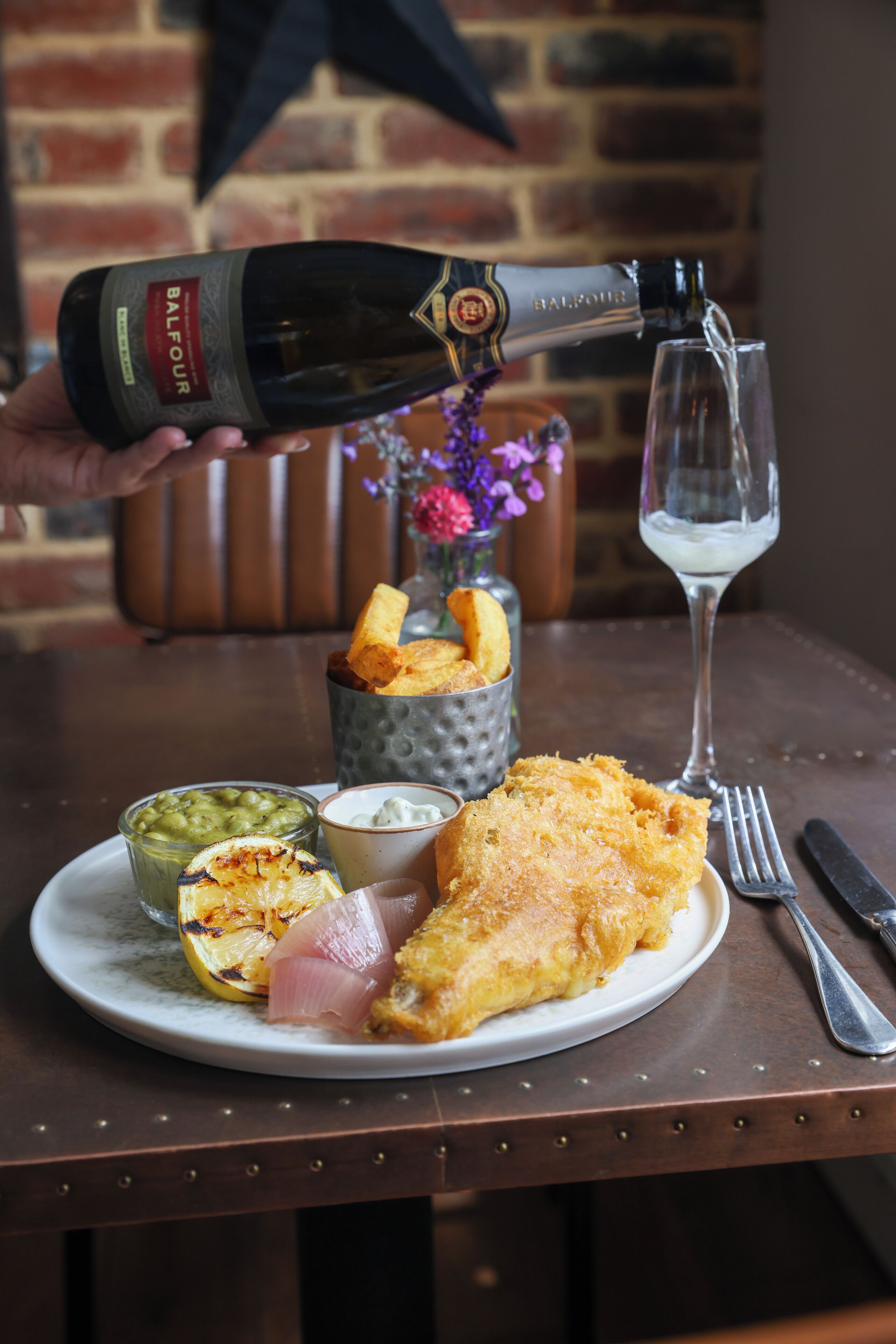 A plate with fish and chips, a glass of water, and a bottle of Balfour wine being poured into a glass.