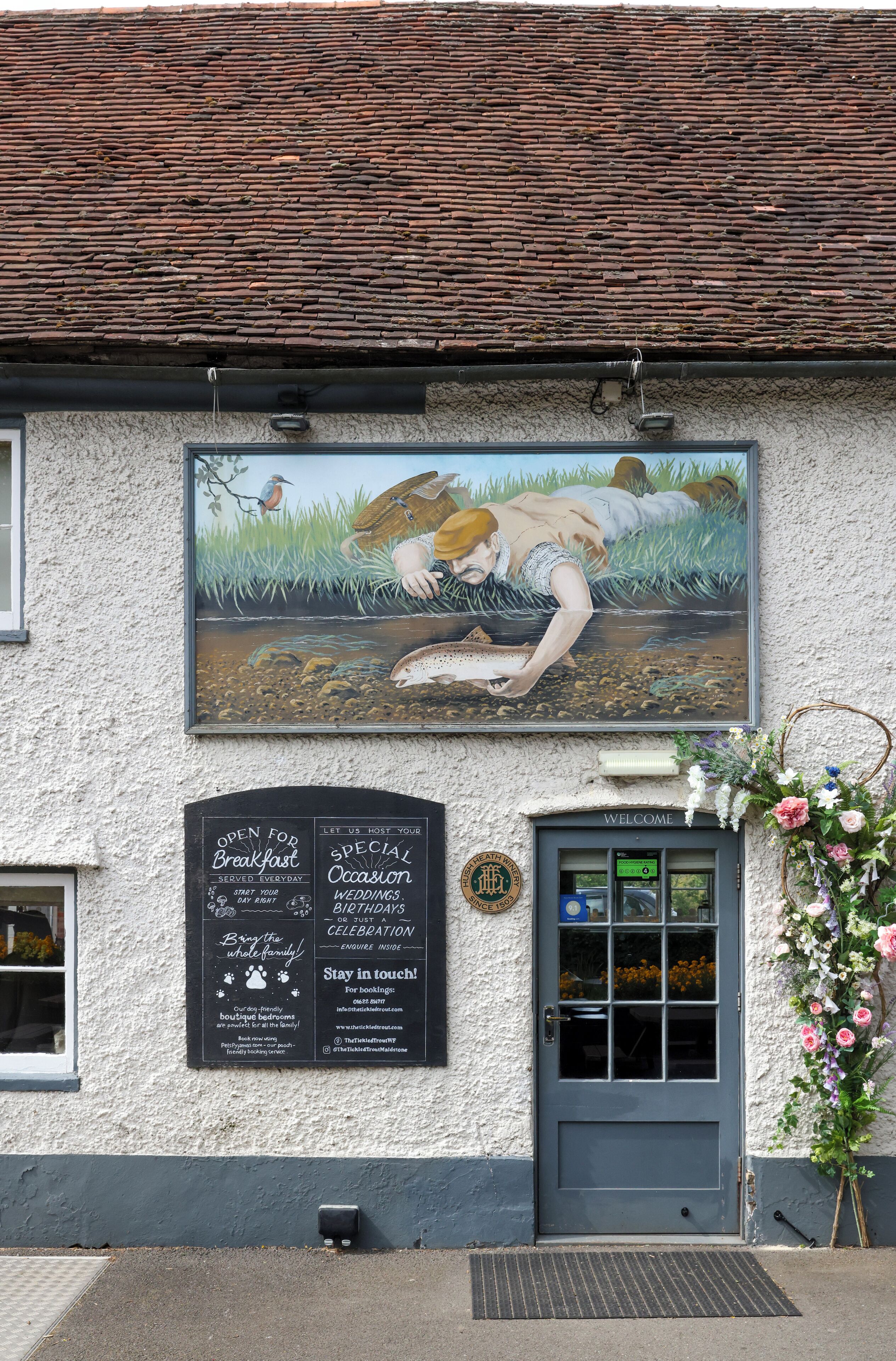 Un bâtiment orné d’une fresque représentant une femme pêchant, une porte avec une pancarte « Bienvenue » et un tableau noir affichant des informations sur les événements.