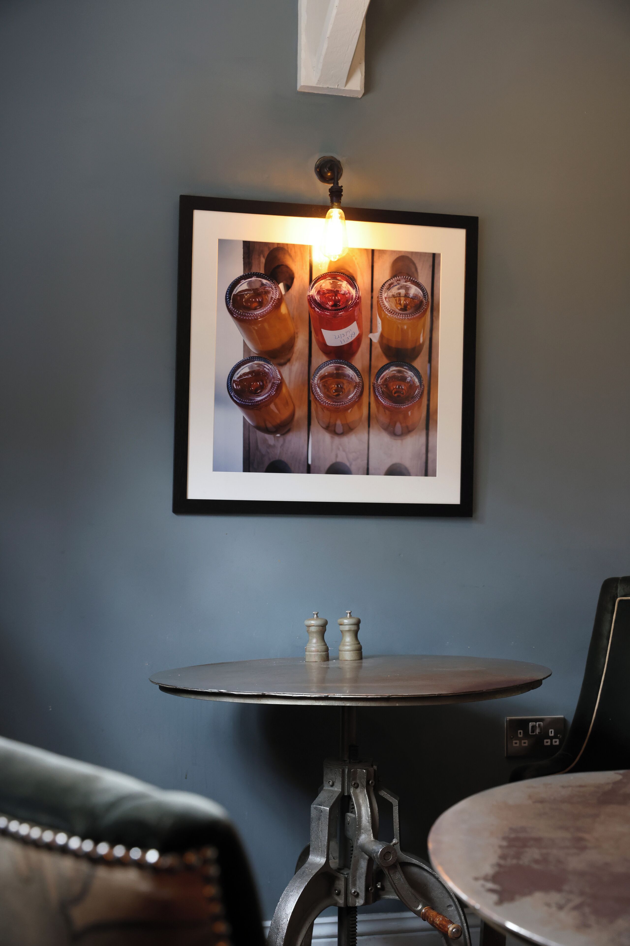 A framed photograph of glasses on a wall, a small round table with a metal base, and a chair with a patterned cushion.