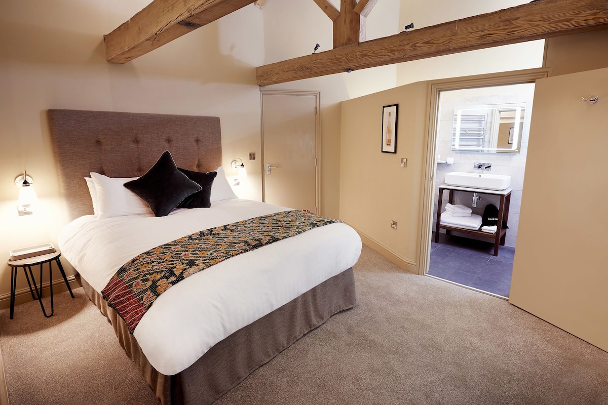 A bedroom with a large bed, a wooden ceiling, a small stool, and a bathroom visible through an open door.