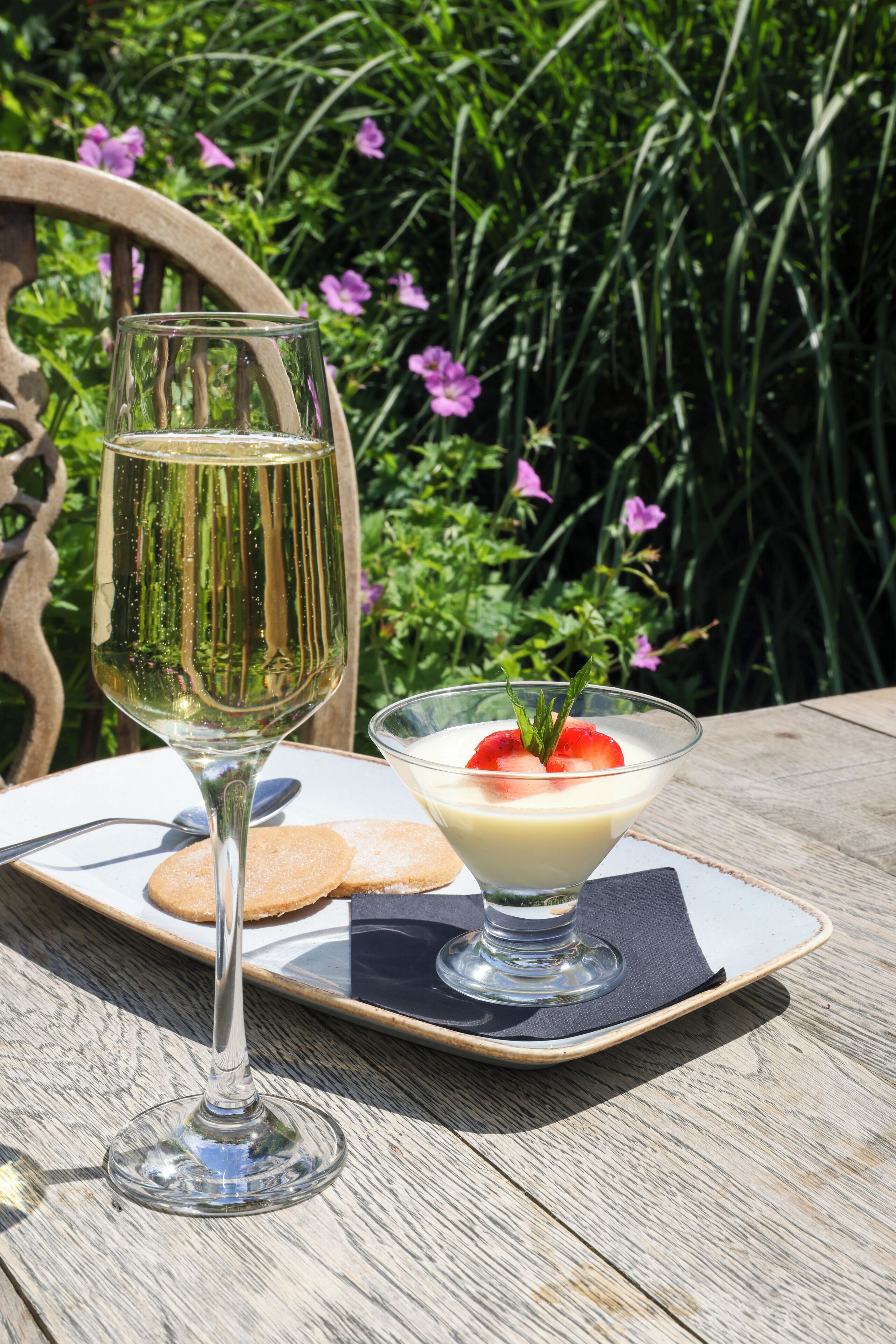 Una copa de vino blanco sobre una mesa de madera, con un postre en un bol de vidrio y una galleta.