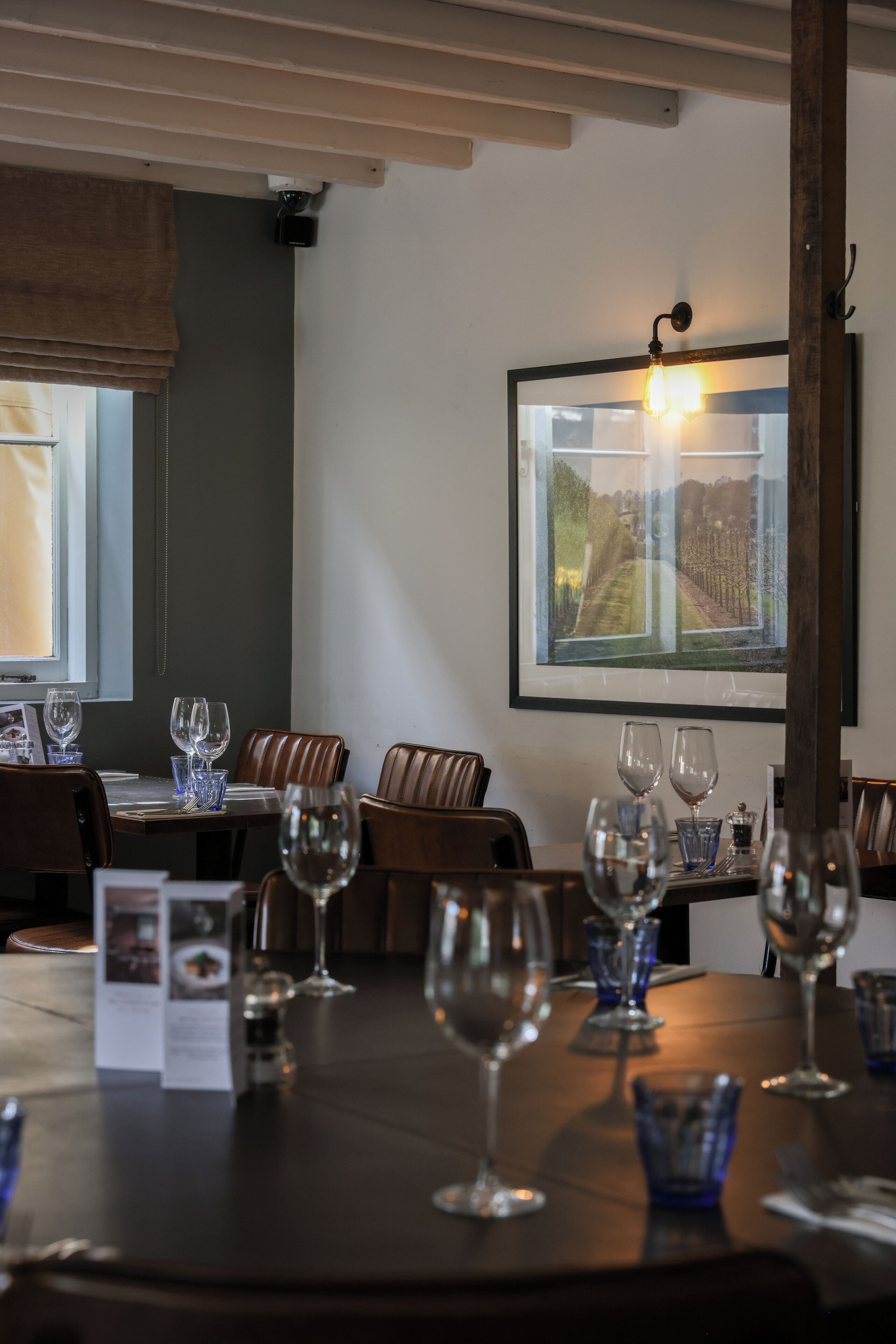 A restaurant with a table set for dining, a framed picture on the wall, and a window with a view of a vineyard.