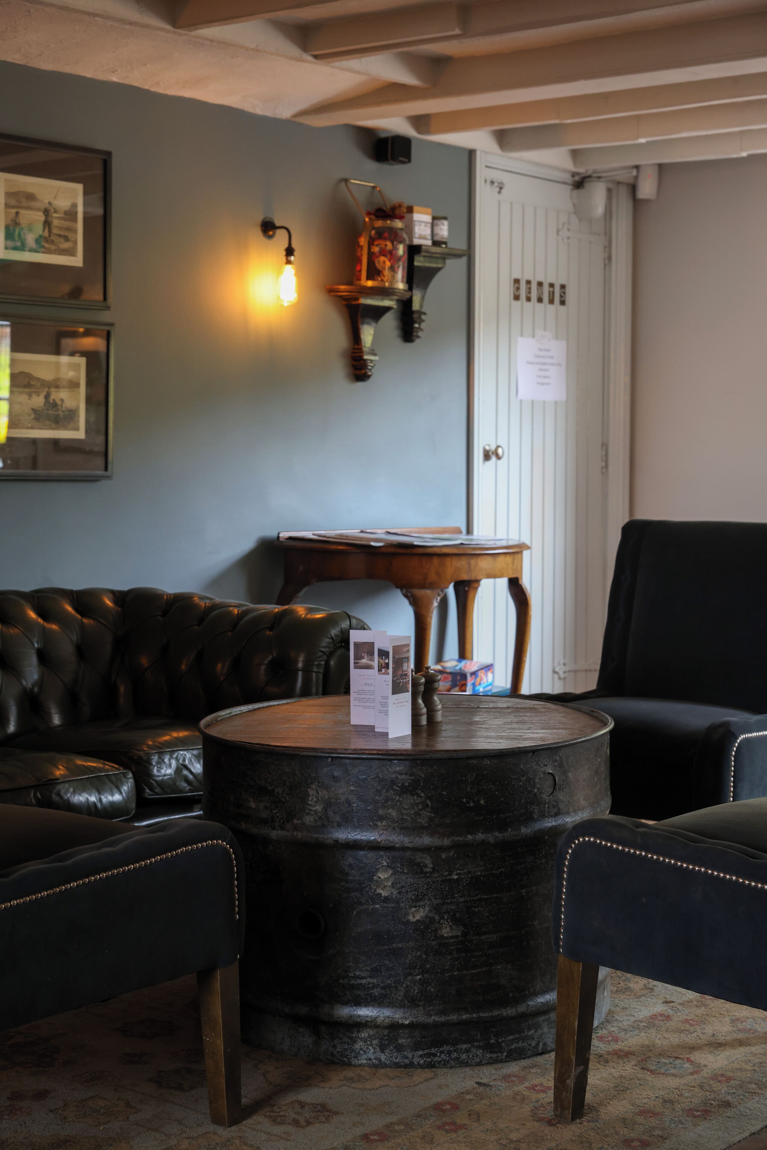 A room with a leather sofa, a round coffee table, and a black armchair. There is a wooden side table with a lamp and a shelf with decorative items. A door with a white frame and a notice on it is visible in the background.