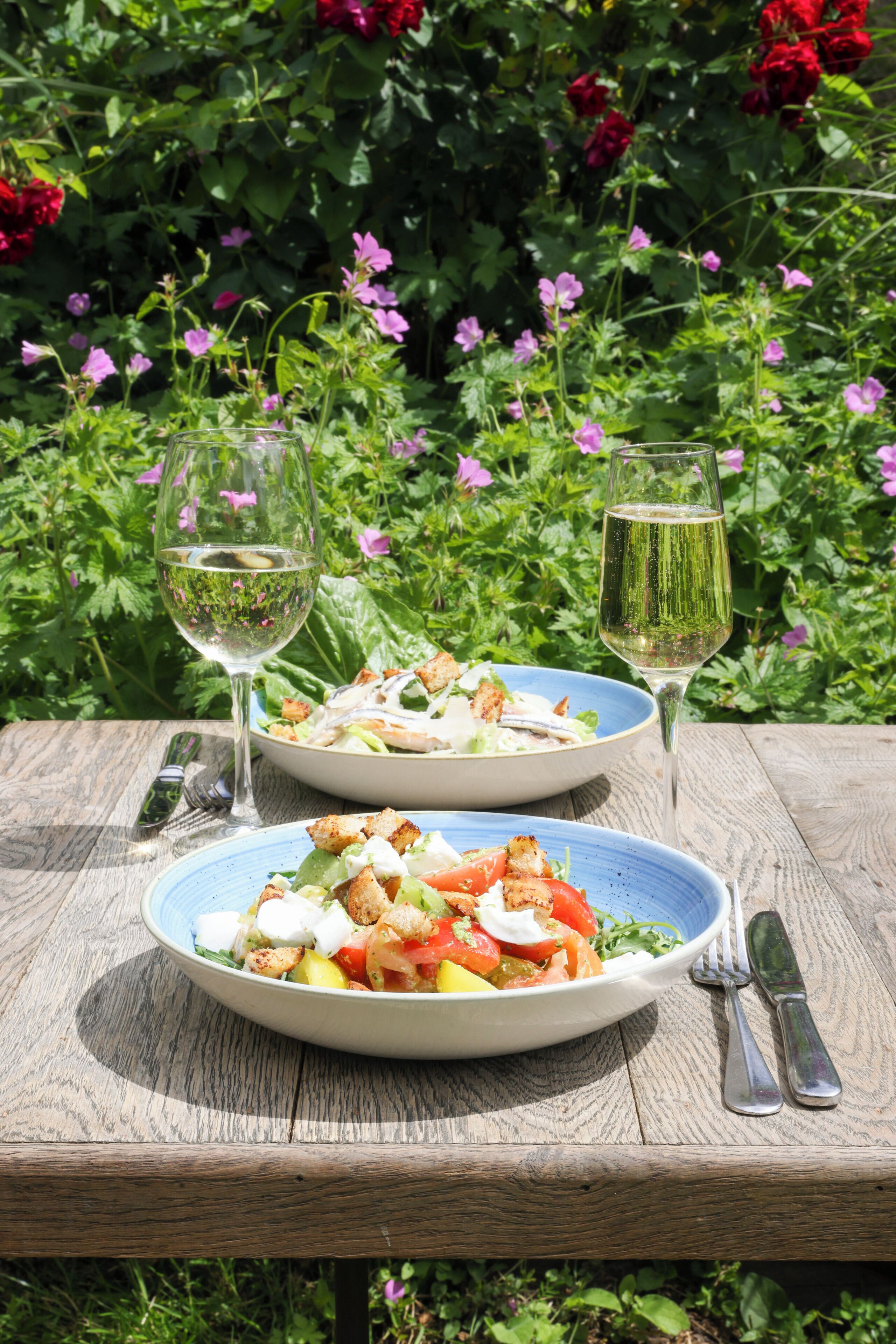 Two plates of salad and two glasses of white wine on a wooden table.