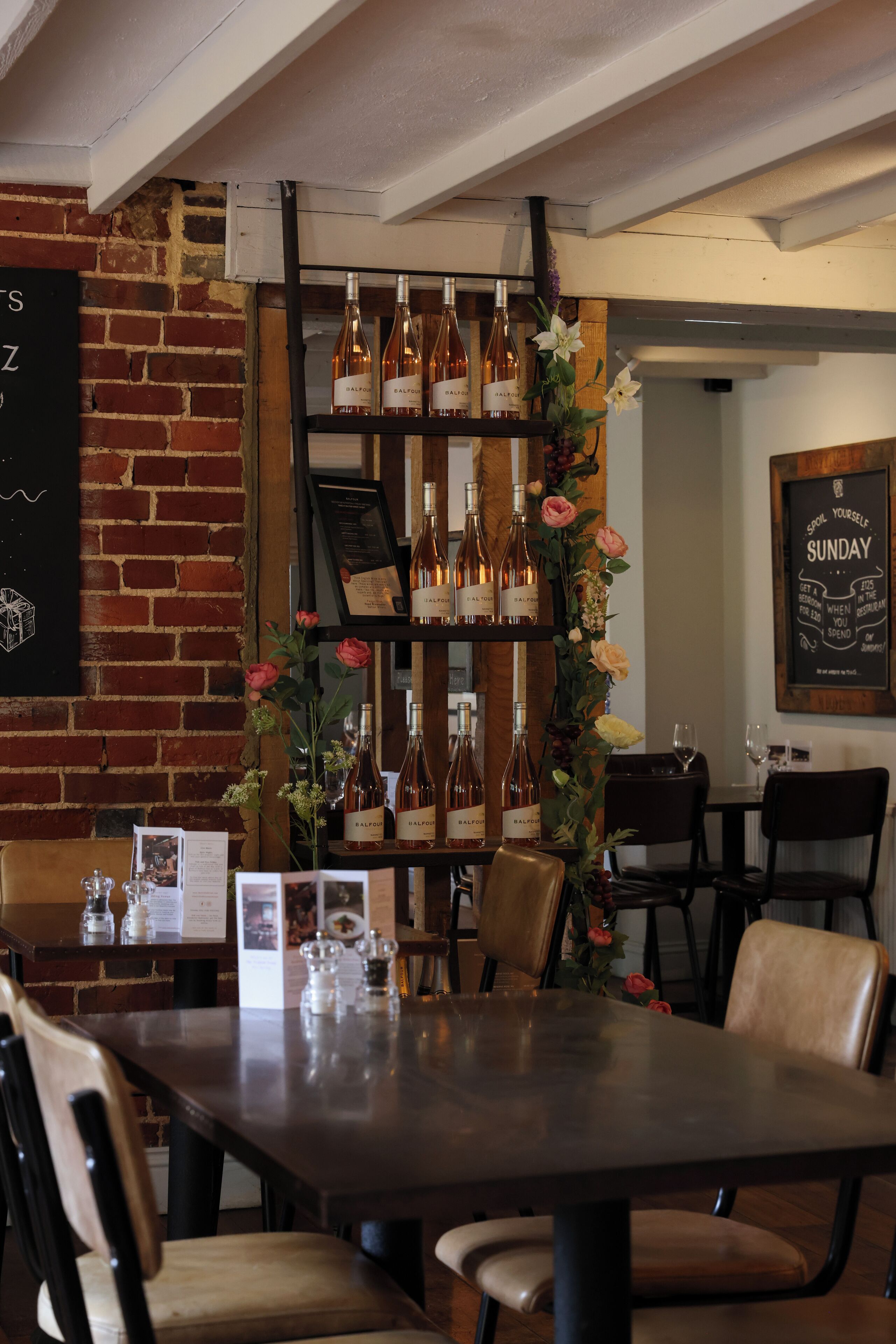 Een restaurantinterieur met bakstenen muren, houten tafels en stoelen. Er staan wijnflessen op planken en een bloemstuk staat er.