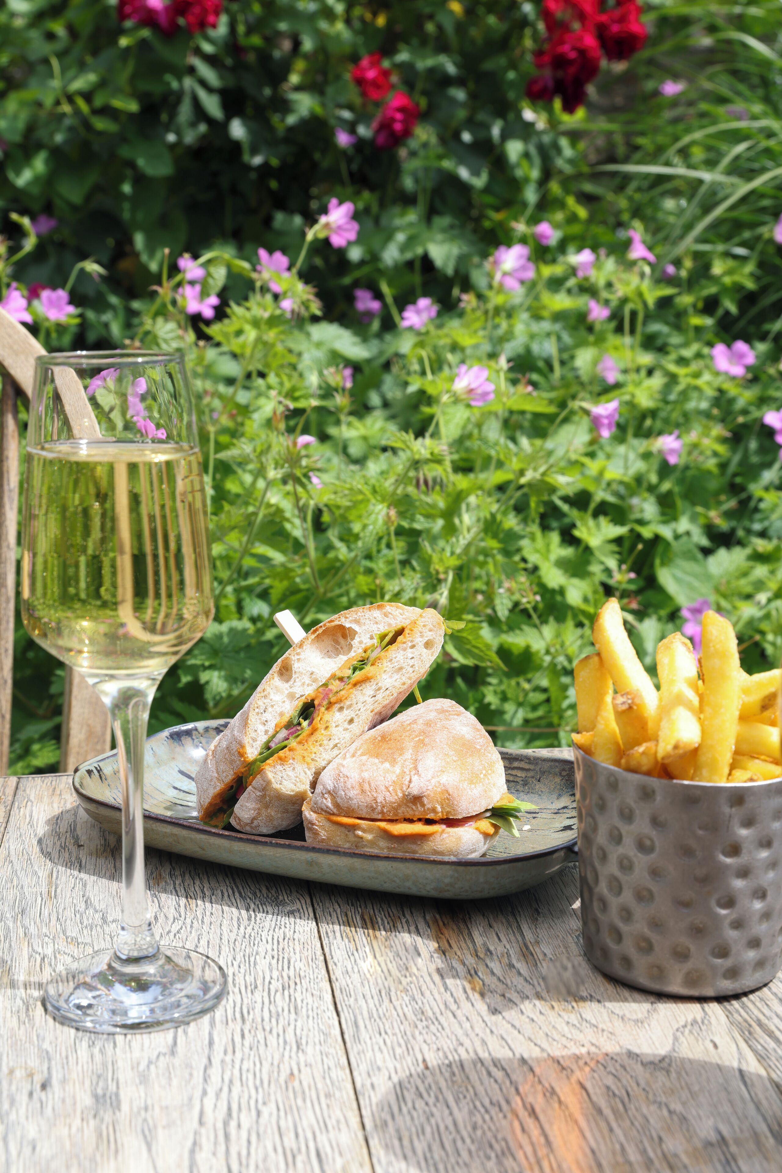 A table with a plate of sandwiches, a glass of white wine, and a cup of fries.