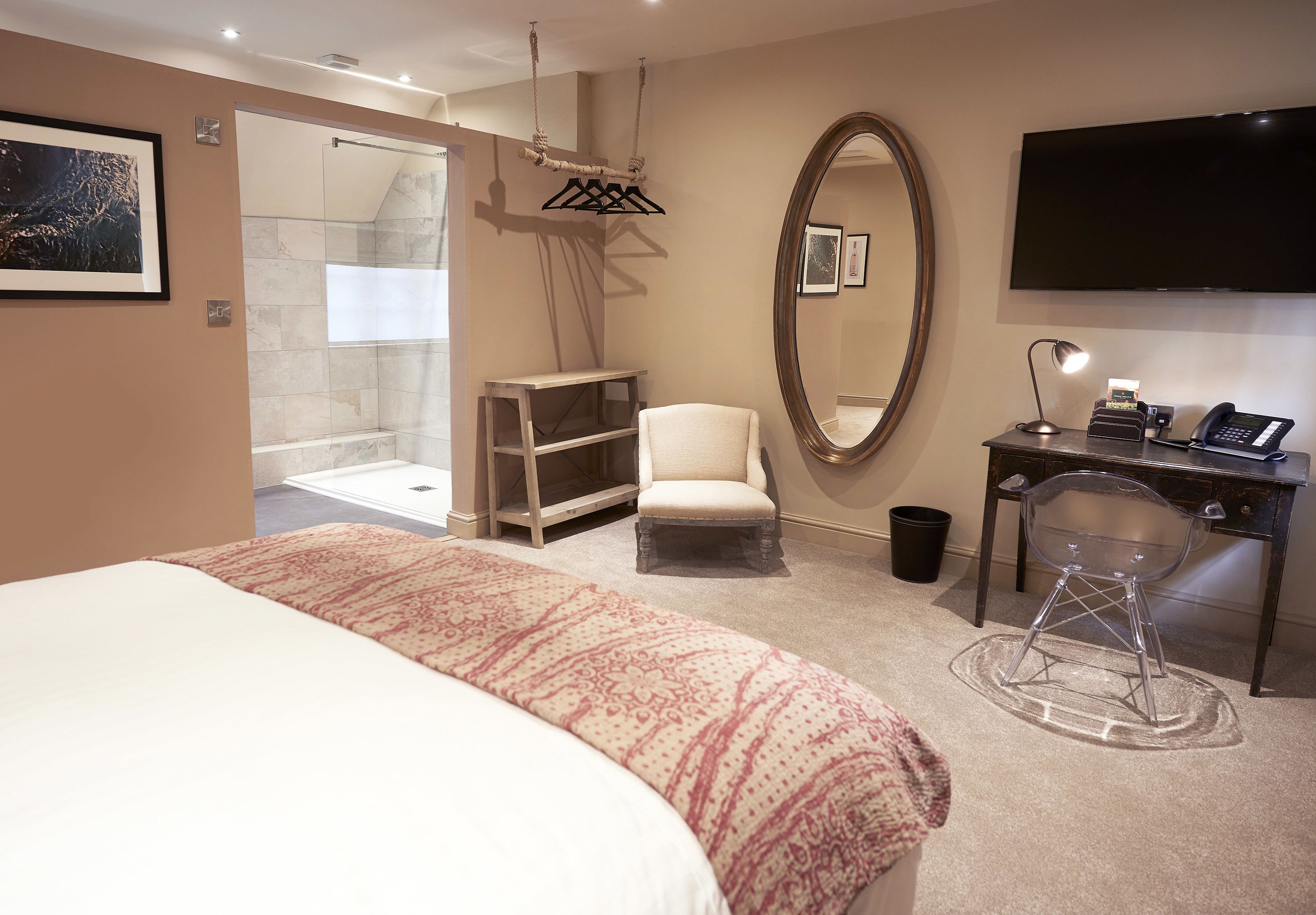A modern hotel room with a large bed, a sitting area with a chair, a desk with a phone, a mirror, a TV, and a shower area.