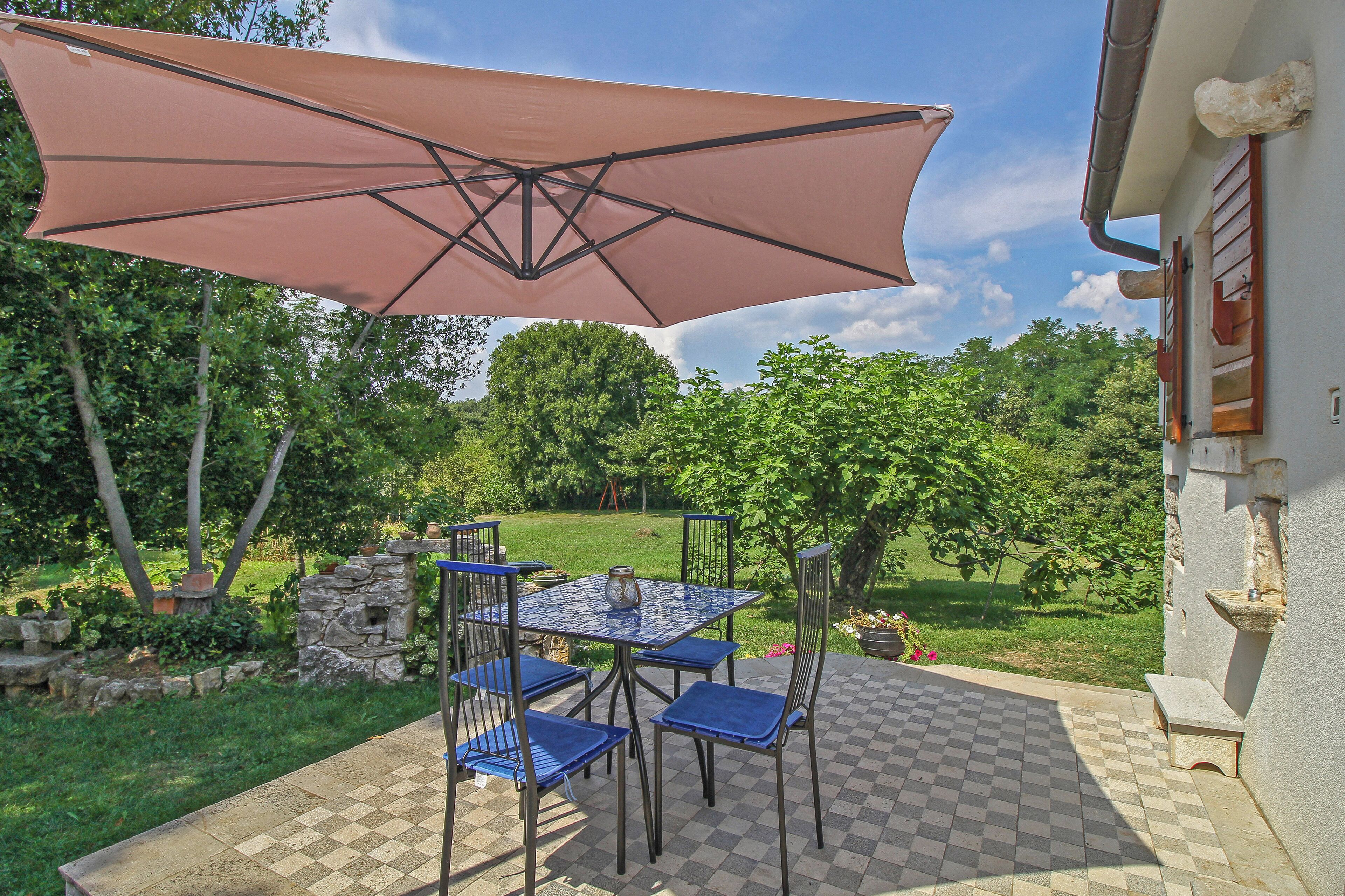 An outdoor patio with a table and chairs, a large umbrella, and a view of a garden.