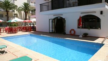A swimming pool with red chairs and umbrellas, a lifebuoy, and a building with a balcony.