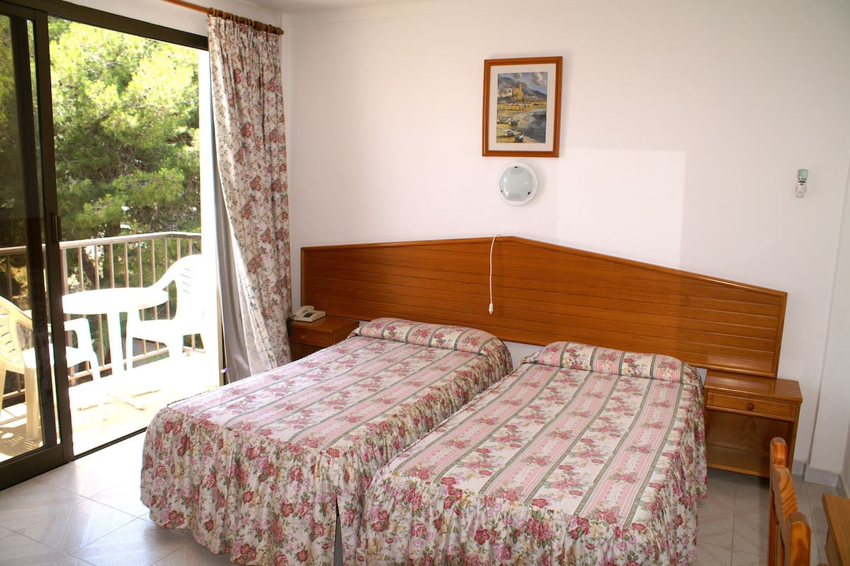 Ein Schlafzimmer mit Holzkopfteil, zwei Betten mit floralem Bettzeug, einem Nachttisch, einem Beistelltisch, einem Stuhl und einem Balkon mit Blick auf Bäume.