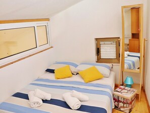 Un dormitorio con cama, almohadas, un espejo, una ventana y una mesita de noche con lámpara.