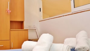 Una cama bien hecha con toallas blancas enrolladas, un armario de madera y una mesita con una silla.