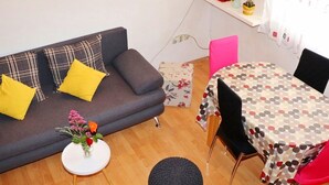 Una sala de estar con un sofá gris, almohadas amarillas, una mesita blanca con un florero, un tapete redondo negro y una mesa de comedor con mantel de lunares.