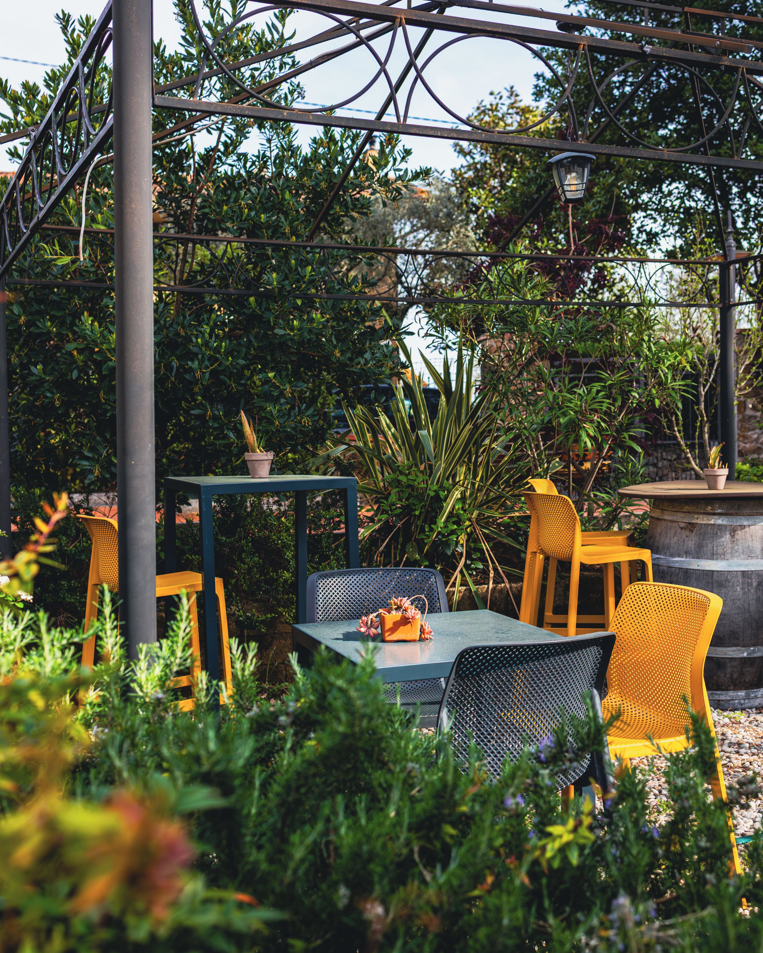 An outdoor seating area with metal chairs and a table, surrounded by greenery and a pergola structure.