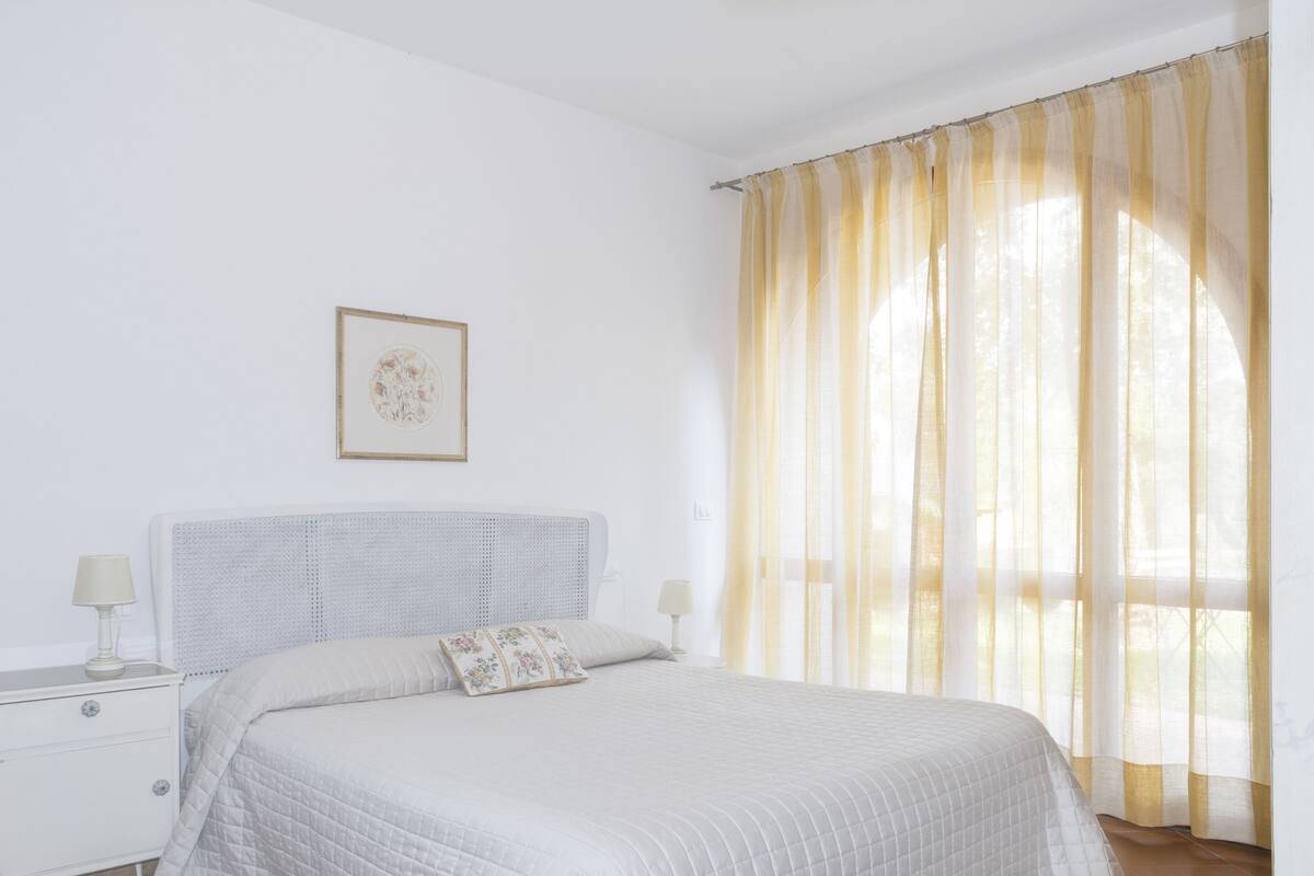 A bedroom with a bed, nightstands, a framed picture on the wall, and sheer curtains.
