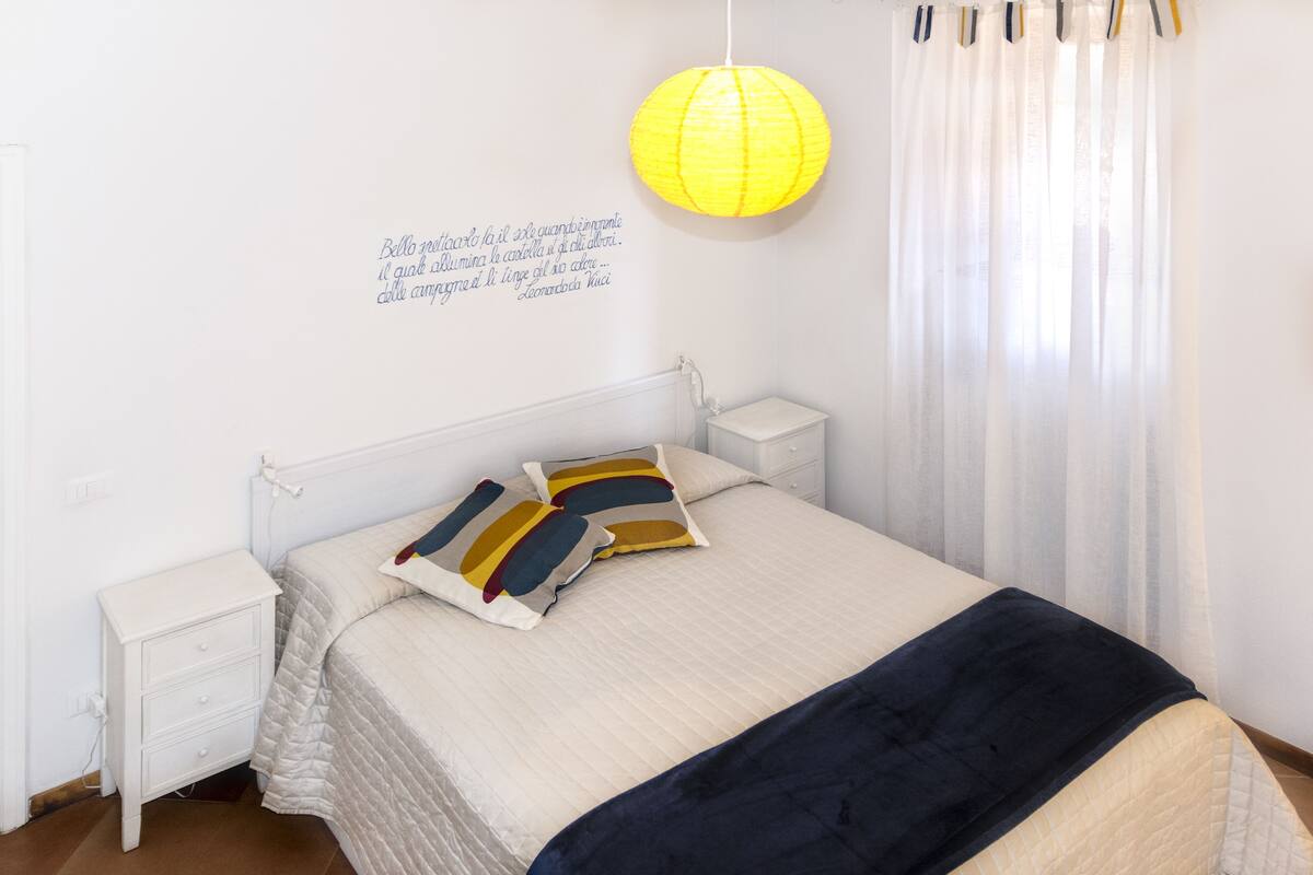 A neatly made bed with two pillows, a bedside table, a wall-mounted lamp, and a quote on the wall.