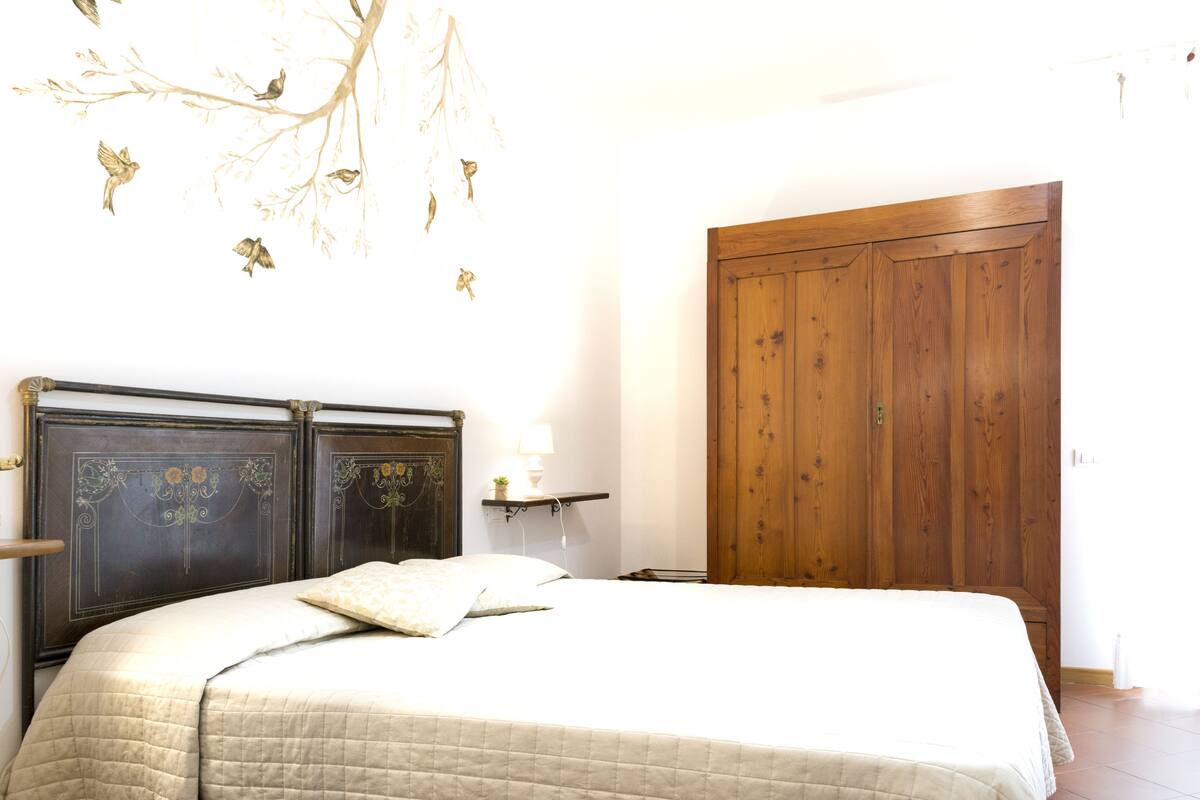 A bedroom with a bed, a wooden wardrobe, a wall-mounted shelf with a lamp, and a wall decoration of birds.