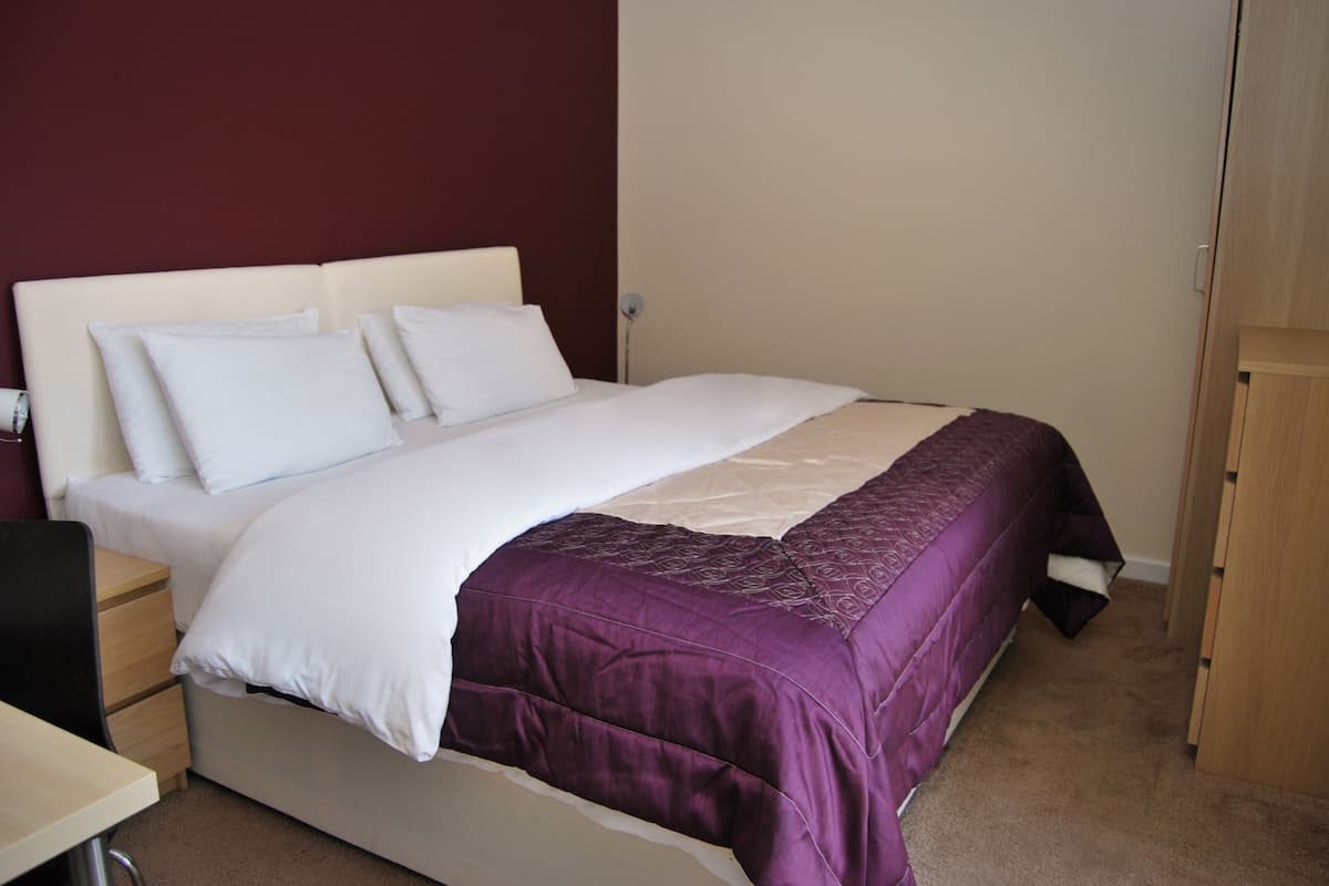 A neatly made bed with white and purple bedding, a wooden nightstand, and a desk with a chair.