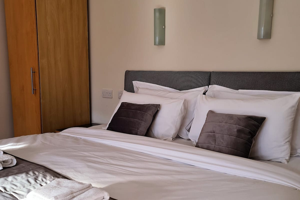A neatly made bed with white linens and two pillows, a wooden wardrobe, and two wall-mounted light fixtures.