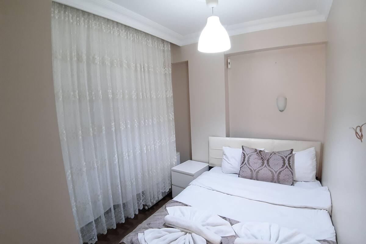 A hotel room with a bed, a nightstand, a lamp, and a window with sheer curtains.