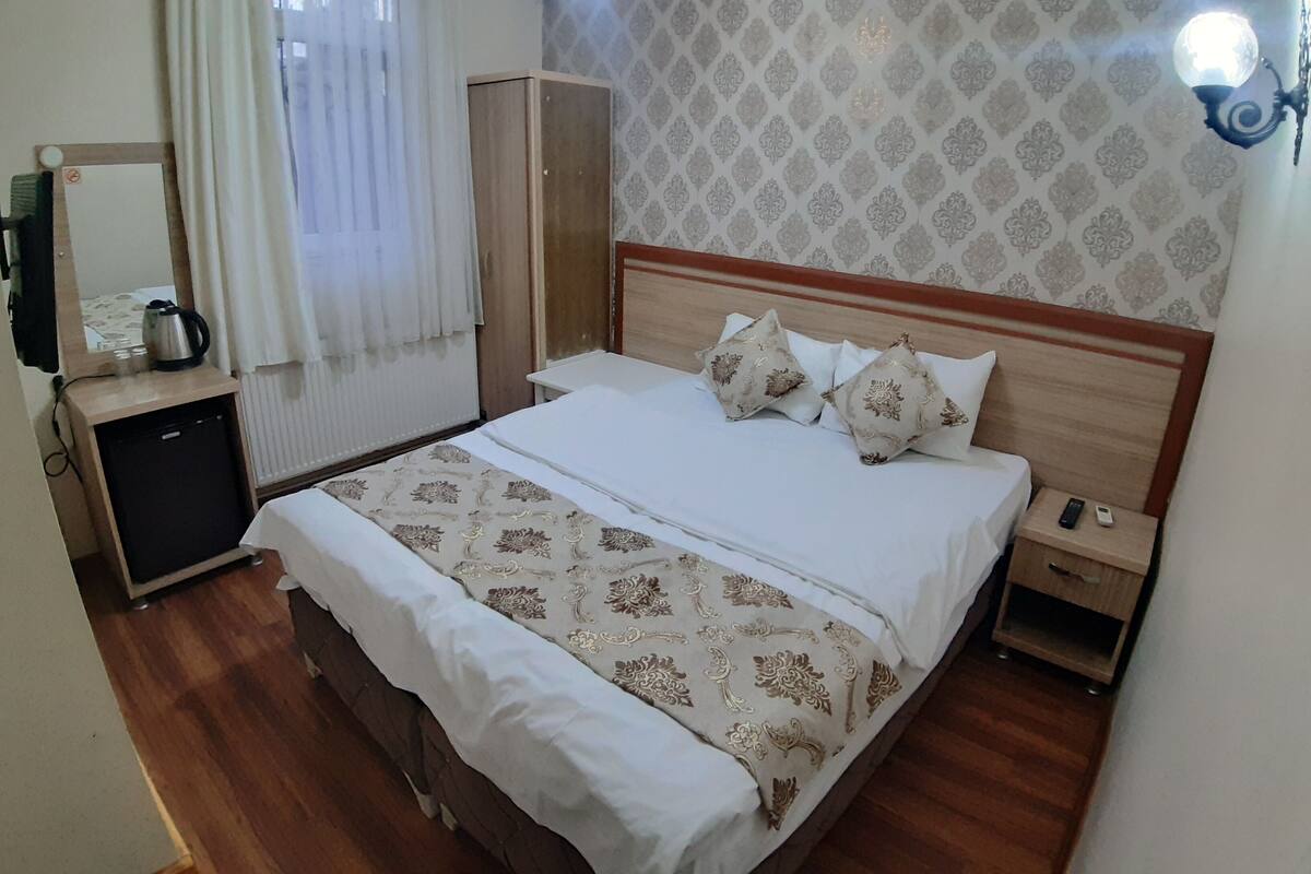 A neatly arranged hotel room with a bed, bedside tables, a wardrobe, a mirror, and a small desk with a kettle.