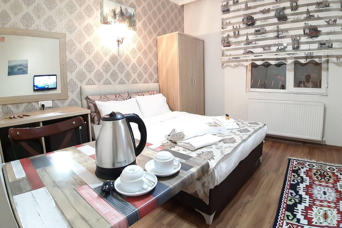 A neatly arranged hotel room with a bed, a desk, a kettle, and a table set with cups and a plate.