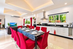 A modern kitchen with white cabinets, a large window with a view of greenery, and a dining area with red chairs and a blue tablecloth.