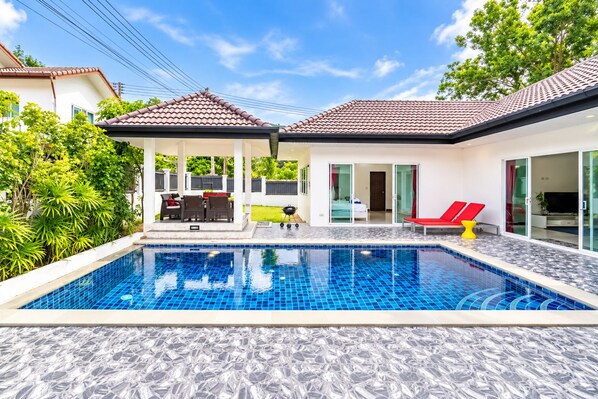 A swimming pool with a tiled deck, a covered patio with furniture, and a glass door leading to a room. - Chalong 4BR Private Pool Villa | Near Big Buddha | Sleeps 10 (Tambon Chalong)