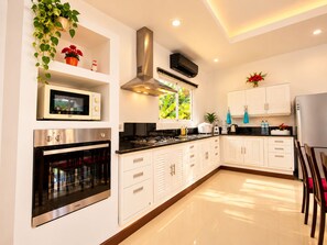 A modern kitchen with built-in appliances, white cabinetry, and a window with a view of greenery. - Chalong 4BR Private Pool Villa | Near Big Buddha | Sleeps 10 (Tambon Chalong)