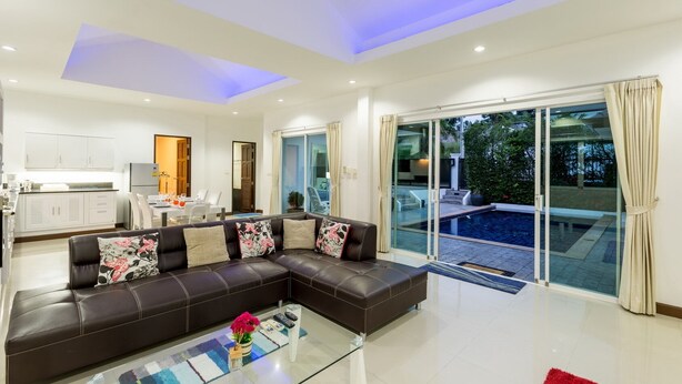 A modern living room with a sectional sofa, a glass coffee table, and a view of a swimming pool through large sliding glass doors. - Chalong 4BR Private Pool Villa | Near Big Buddha | Sleeps 10 (Tambon Chalong)