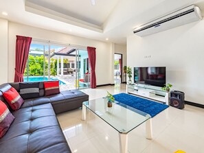 A modern living room with a glass coffee table, a leather sofa, a flat-screen TV, and a pool view. - Chalong 4BR Private Pool Villa | Near Big Buddha | Sleeps 10 (Tambon Chalong)