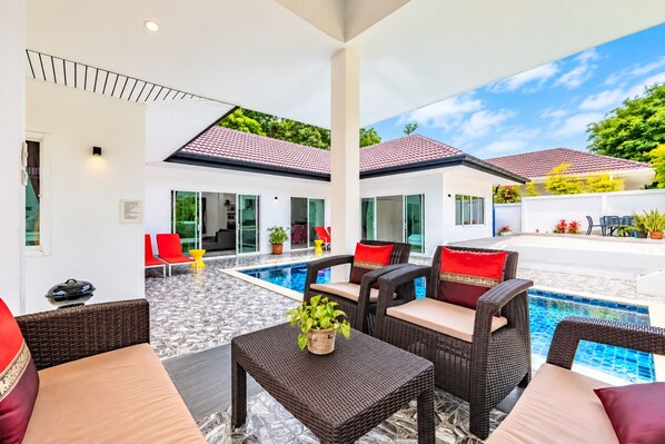 A modern outdoor pool area with wicker furniture and red cushions, surrounded by a white building with glass doors.