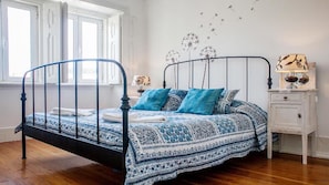 Camera da letto con una struttura del letto in metallo, una coperta a motivi decorativi, un comodino con una lampada e una decorazione murale raffigurante tarassaci.