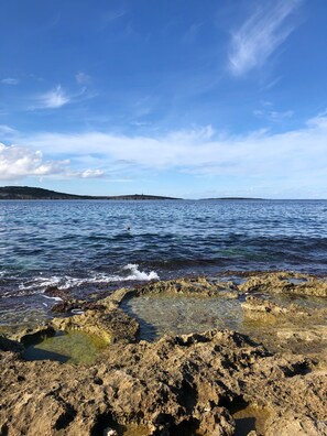 A rocky shoreline with clear water and a distant landmass under a blue sky.
