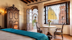 Een slaapkamer met een groot bed, een houten garderobe, een klein tafeltje met een vaas en uitzicht op een klokkentoren door een raam met ijzeren tralies.