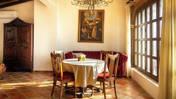 A dining area with a round table, chairs, a chandelier, a painting on the wall, and a window with curtains.